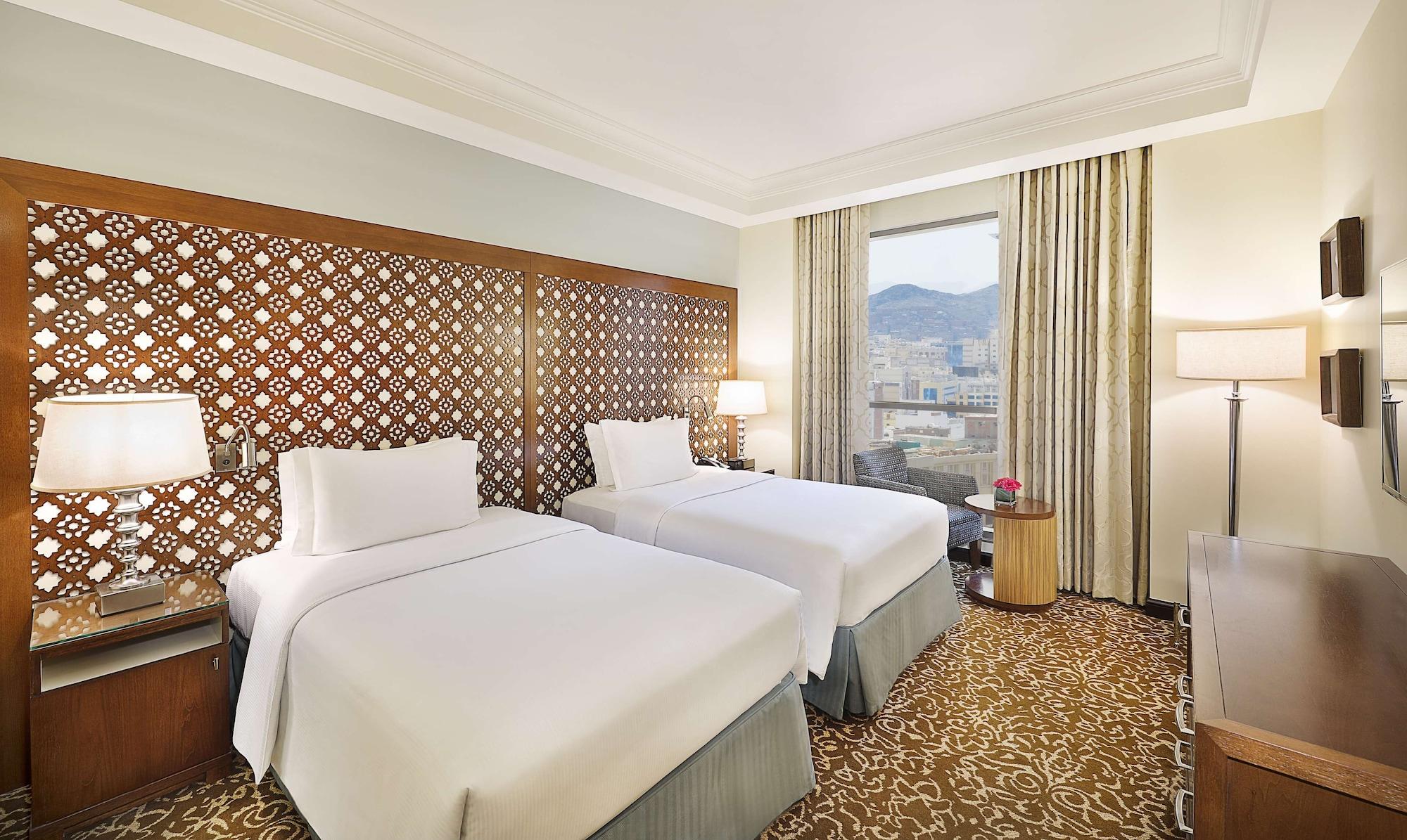 希尔顿套房麦加酒店 Mecca 外观 照片 The photo shows a hotel room featuring two beds with white linens. The beds are positioned against a decorative wooden headboard with an intricate lattice design. There are bedside lamps on either side of the beds. A large window allows natural light