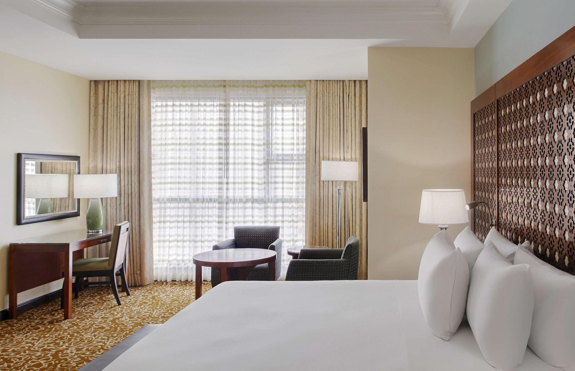 希尔顿套房麦加酒店 Mecca 外观 照片 The photo shows a hotel room with a modern design. In the foreground, there is a large bed with several white pillows. In the background, you can see a window with sheer curtains that allow natural light to fill the room. There are two armchairs and 