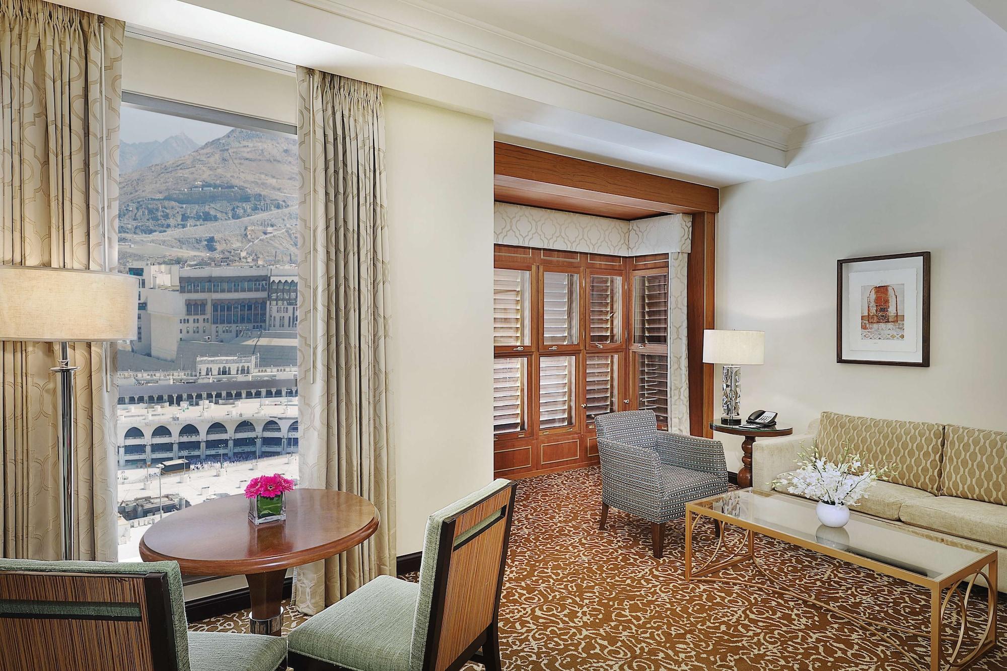 希尔顿套房麦加酒店 Mecca 外观 照片 The photo shows a luxurious hotel room with a welcoming ambiance. There are large windows that provide a view of the outside, suggesting a well-lit space. The room features a seating area with a couch and chairs, alongside a small round dining table.