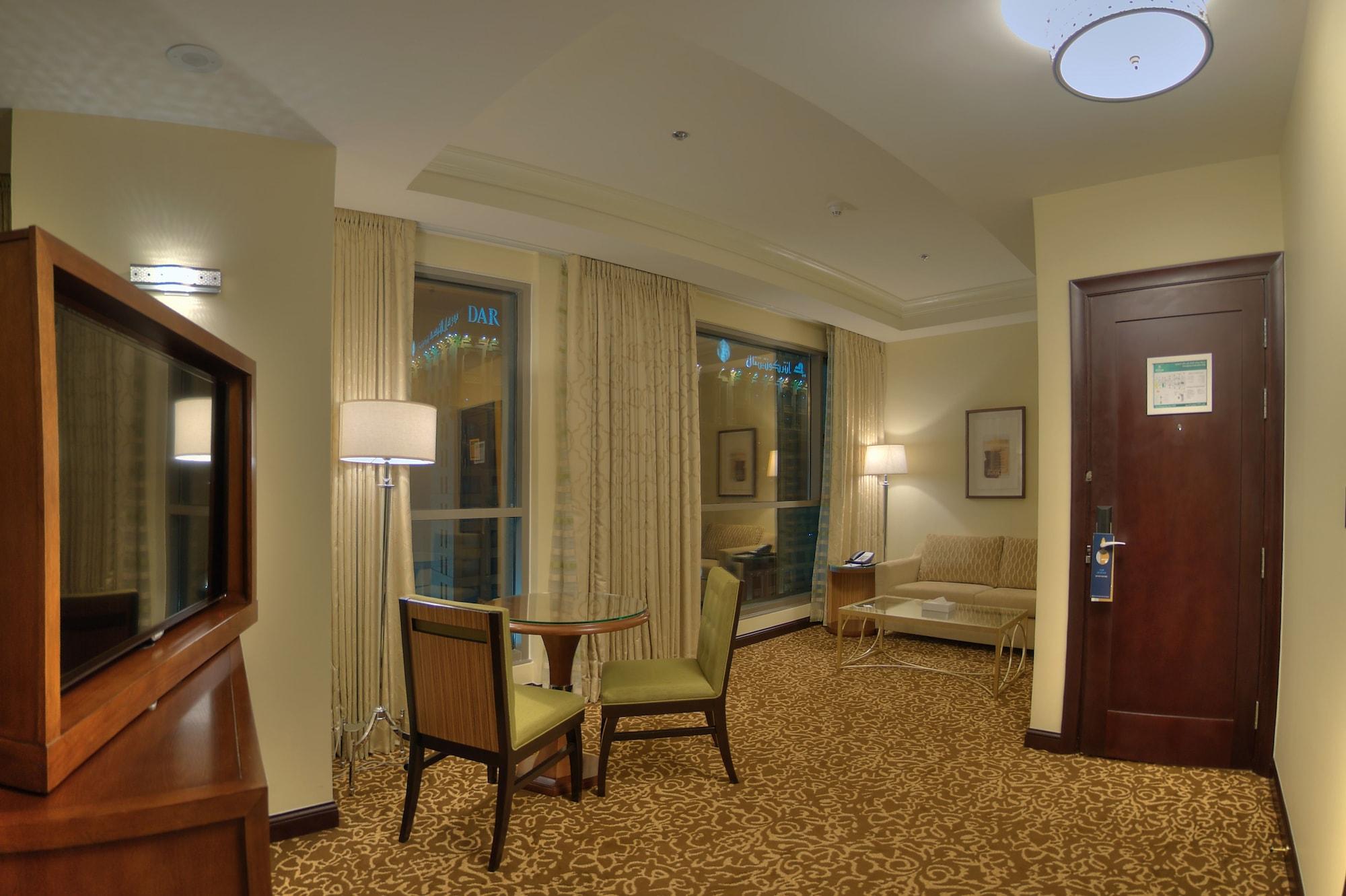 希尔顿套房麦加酒店 Mecca 外观 照片 The photo shows a cozy hotel room or suite that features a small seating area. There is a table with two chairs, suggesting a space for dining or working. A comfortable-looking sofa is positioned against the wall, next to a window covered with light-