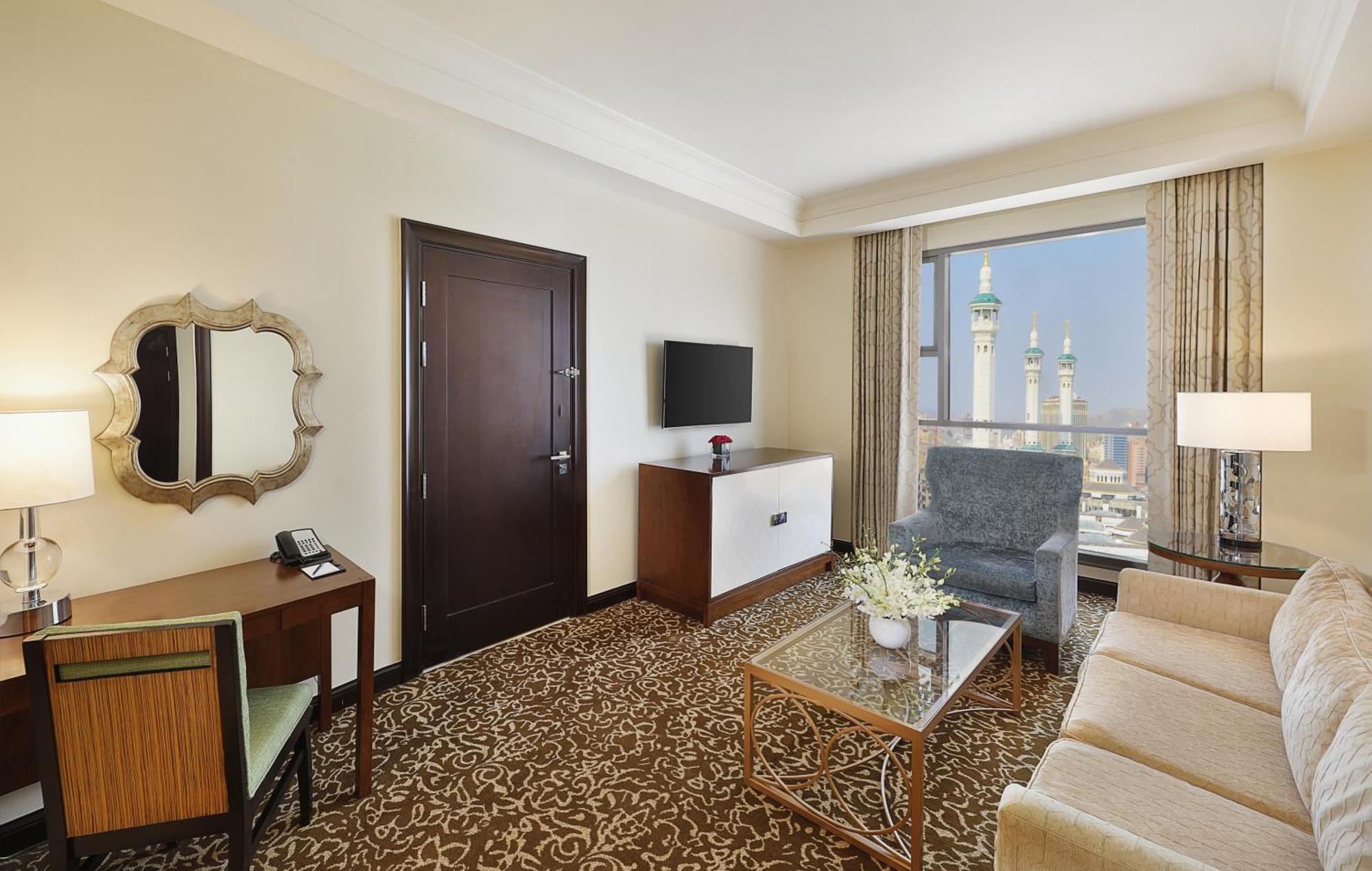 希尔顿套房麦加酒店 Mecca 外观 照片 The photo shows a hotel room with a modern and elegant design. It features a cozy seating area with a beige sofa and a blue armchair, complemented by a glass coffee table. There is a wooden desk with a mirror and a phone, and a television mounted on 