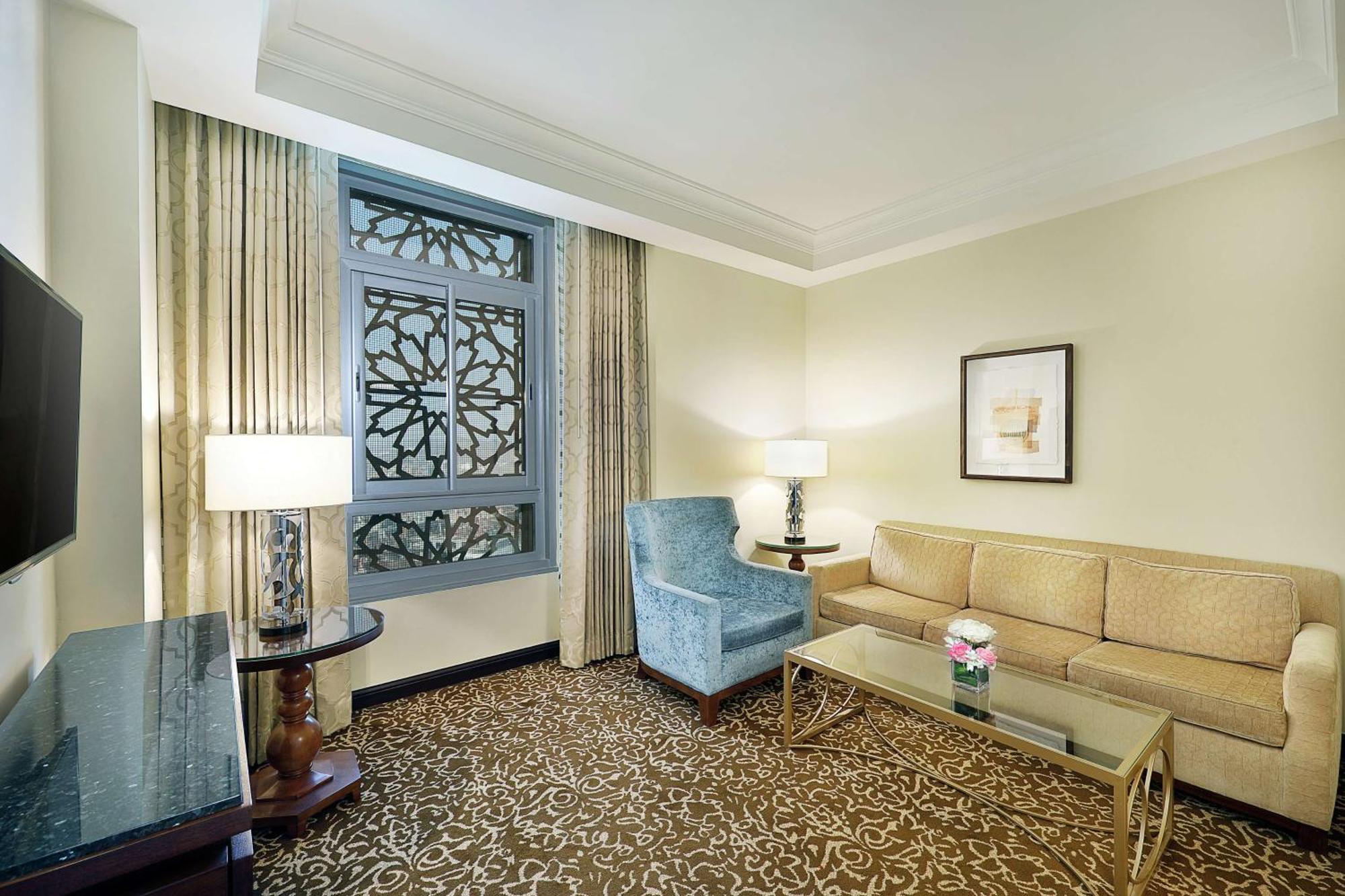 希尔顿套房麦加酒店 Mecca 外观 照片 The photo shows a well-decorated hotel room or suite. It features a light-colored wall with a decorative window that has an intricate design. There are two windows adorned with curtains. The furniture includes a beige couch, a blue upholstered armcha