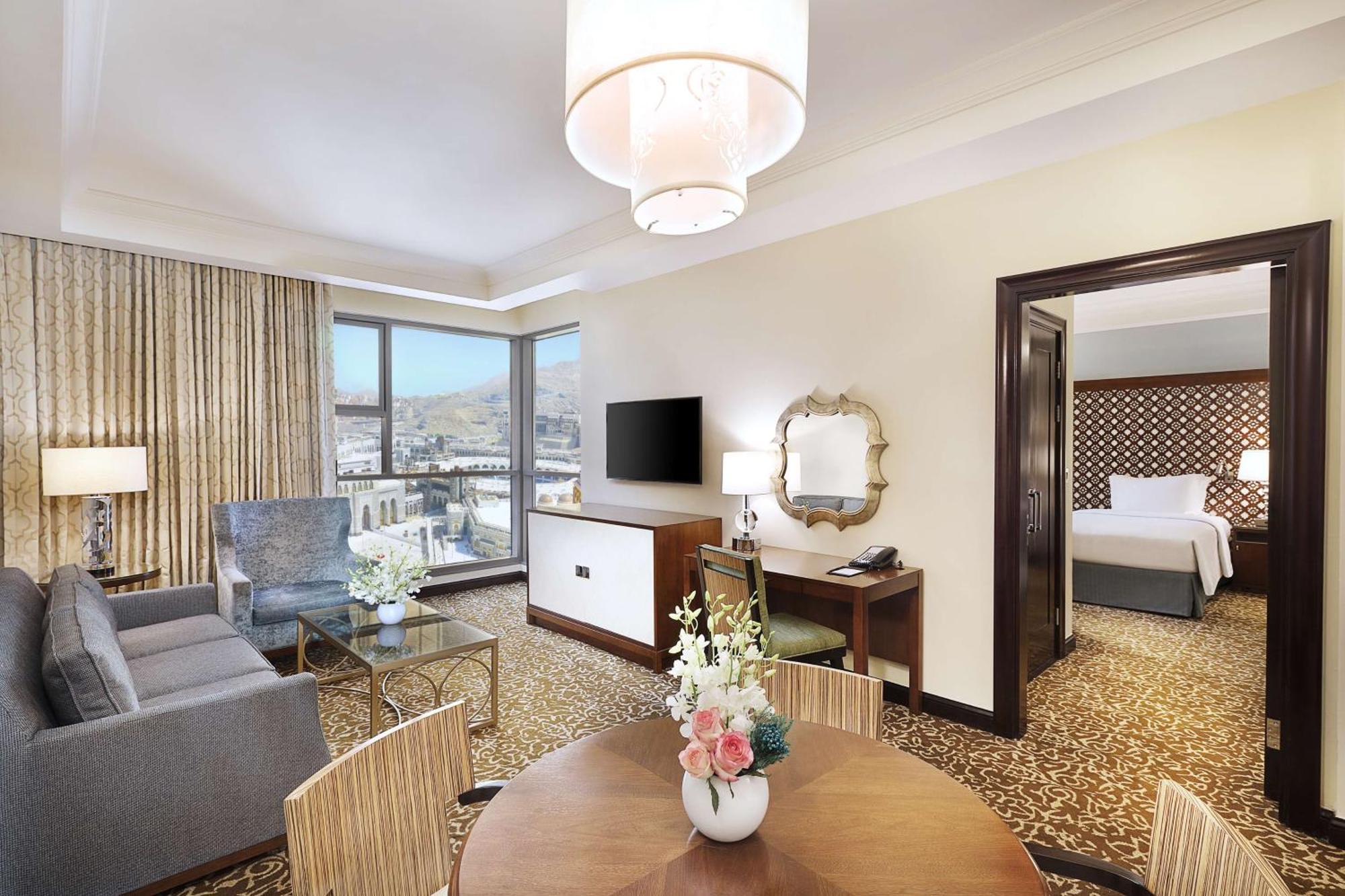 希尔顿套房麦加酒店 Mecca 外观 照片 The image shows a modern hotel room suite. It features a living area with a comfortable gray sofa and a decorative armchair, alongside a round dining table with chairs. There's a flat-screen TV mounted on the wall, and large windows allow natural lig