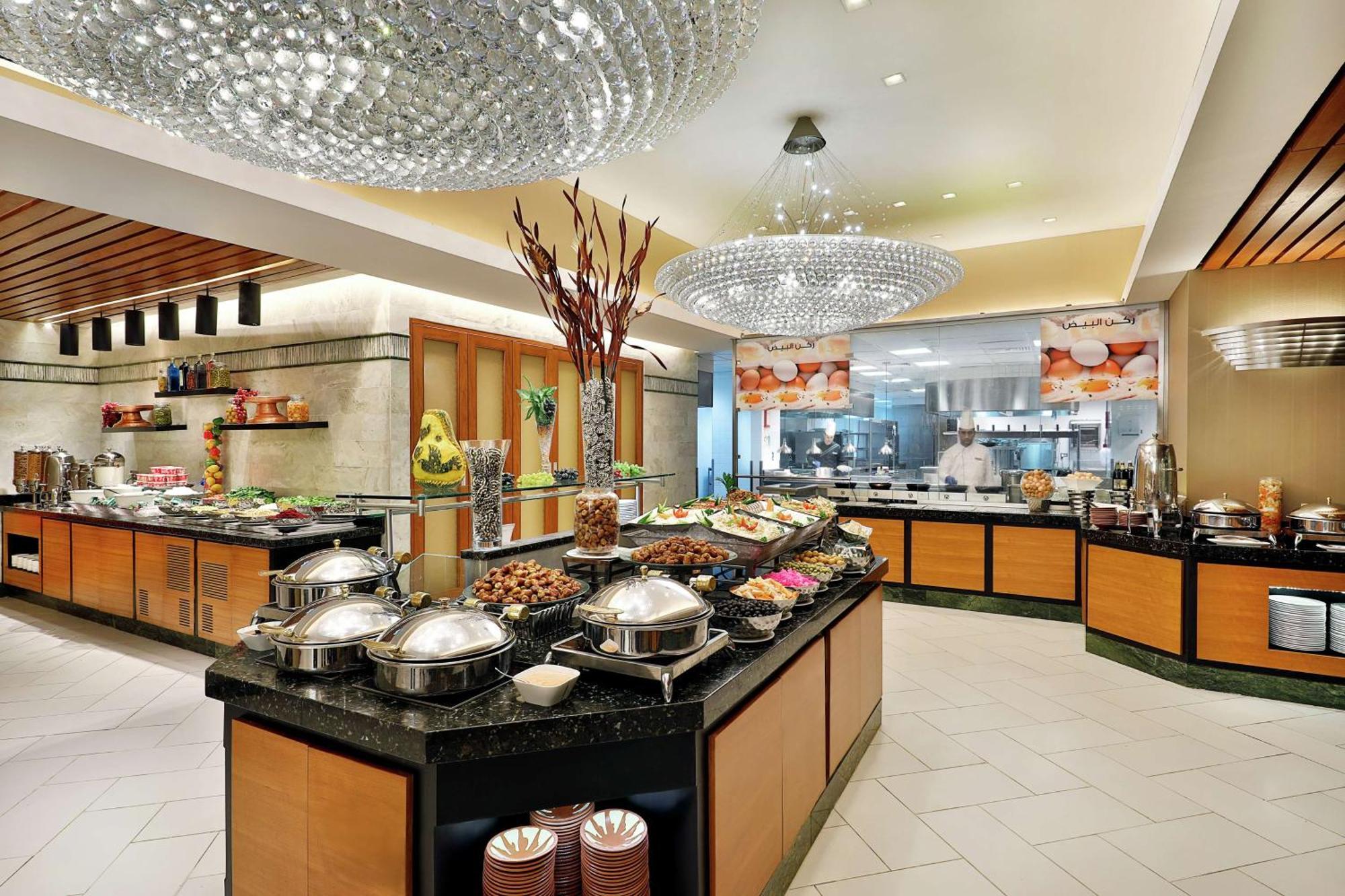 希尔顿套房麦加酒店 Mecca 外观 照片 The photo shows a spacious buffet area in a restaurant or hotel. The design features elegant decor, including bright chandeliers and polished surfaces. There are several buffet tables filled with various food items, displayed attractively. The tables