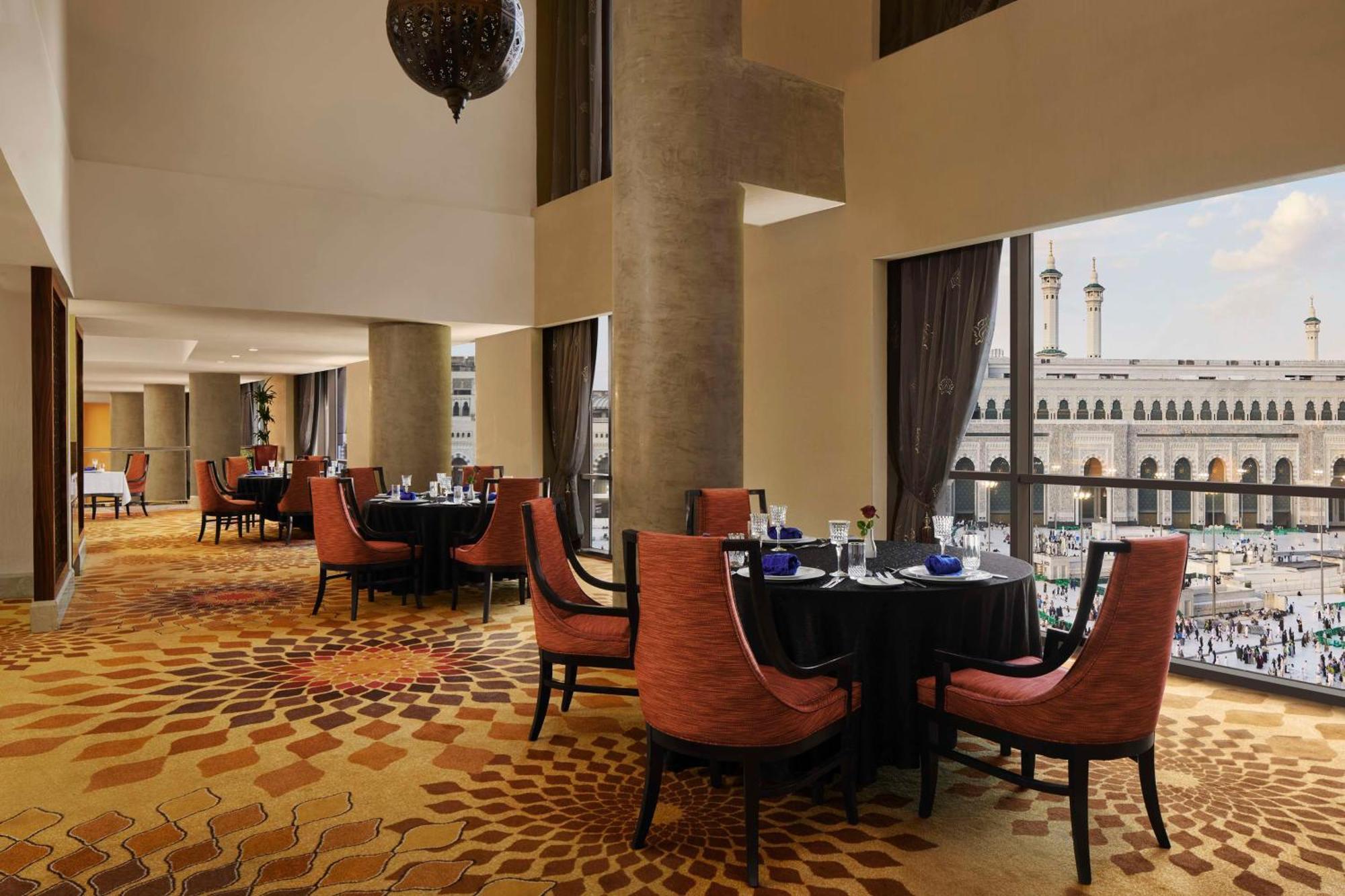 希尔顿套房麦加酒店 Mecca 外观 照片 The photo displays the interior of an elegant restaurant. The design features a spacious layout with high ceilings, large windows offering a view of an exterior area, likely a courtyard or plaza. Inside, there are several round tables set with black 