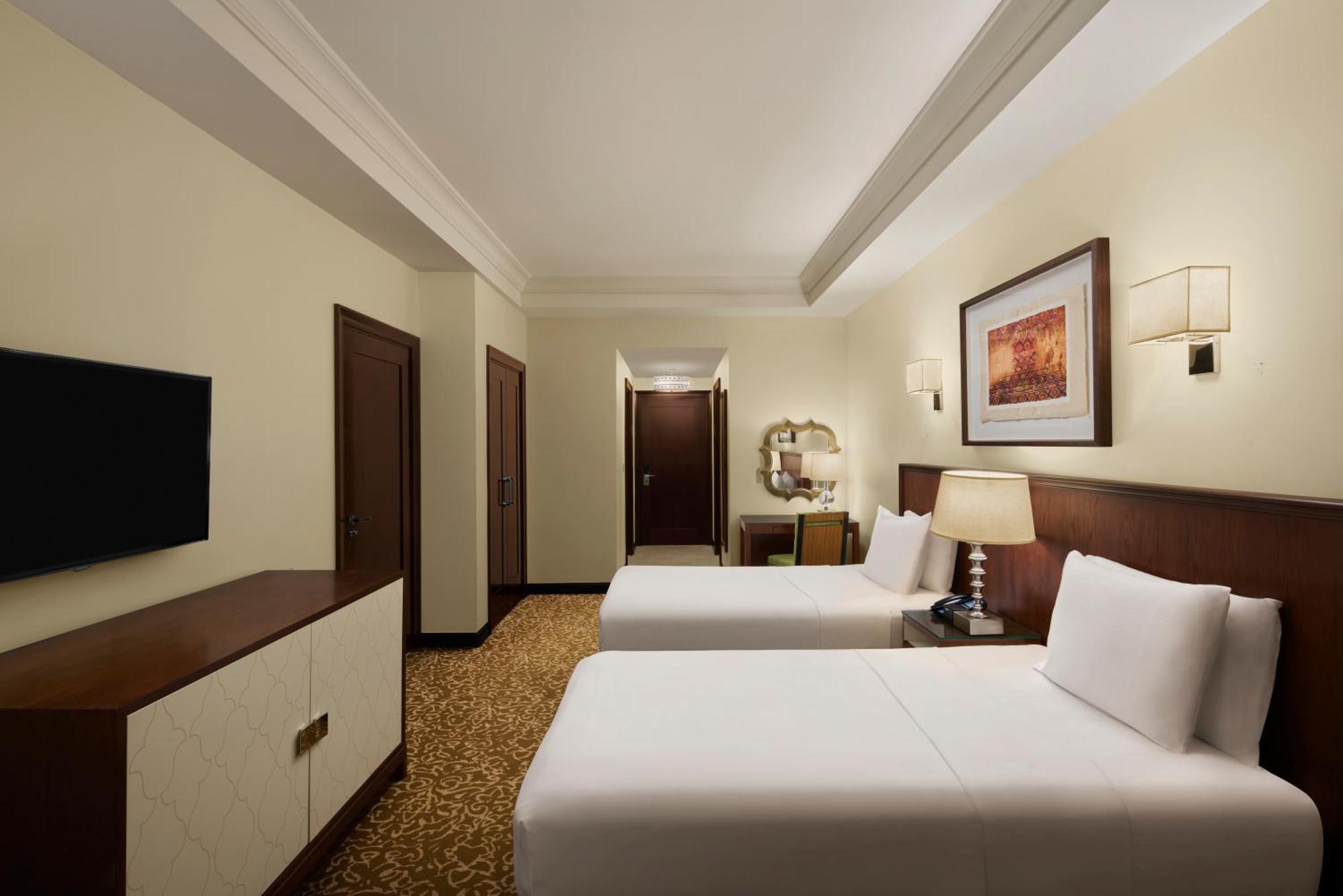 希尔顿套房麦加酒店 Mecca 外观 照片 The photo shows a hotel room that features two beds with white linens. The room has light-colored walls and a warm-colored carpet. There is a wooden dresser with a flat-screen TV mounted on the wall opposite the beds. To the side, there are two doors